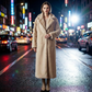 Woman in a long beige coat standing on a city street at night with neon lights.